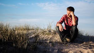 Man in a red plaid shirt sitting thoughtfully on a sandy dune in Australia, surrounded by grass.